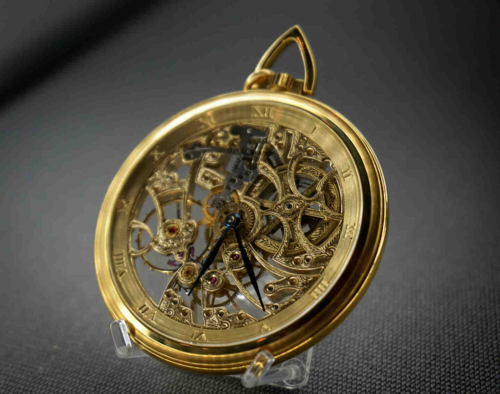 Close-up of an intricately designed gold antique pocket watch with visible mechanical gears and Roman numeral hour markers, displayed on a small clear stand against a dark fabric background.