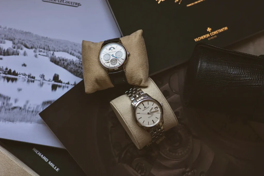 Liquid luxury market display of two high-end watches on suede cushions, with Jaeger-LeCoultre, Vacheron Constantin, and Richard Mille brand materials in the background.