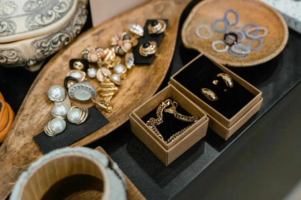 Social media luxury jewelry buying display featuring gold chain bracelets, pearl earrings, and gold rings arranged in kraft boxes and wooden trays on a black surface.