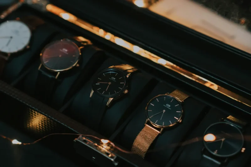 Sell or trade luxury watch collection displayed in a black watch case, featuring multiple Daniel Wellington timepieces with rose gold mesh and leather bands under warm ambient lighting.