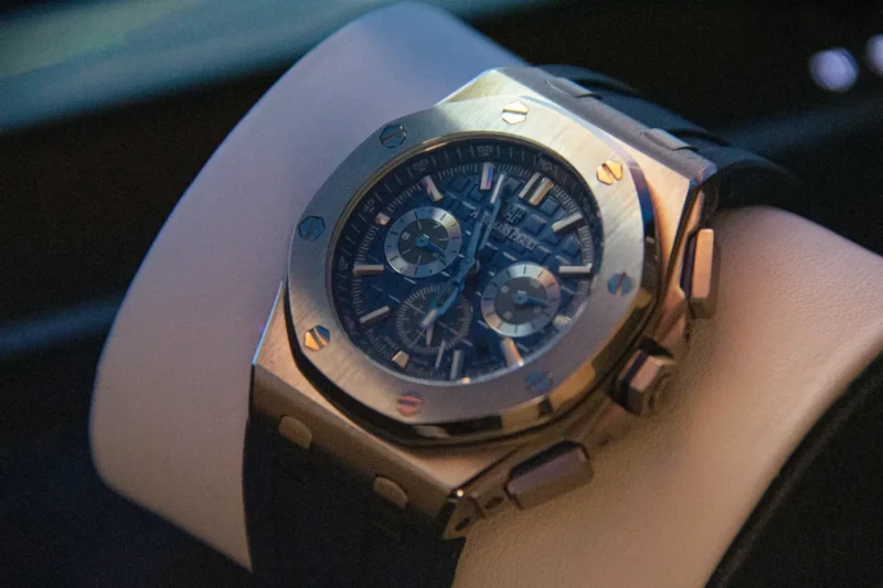 Audemars Piguet Royal Oak chronograph with a blue dial, octagonal steel bezel, and gold screws, displayed on a white cushion in dramatic ambient lighting.